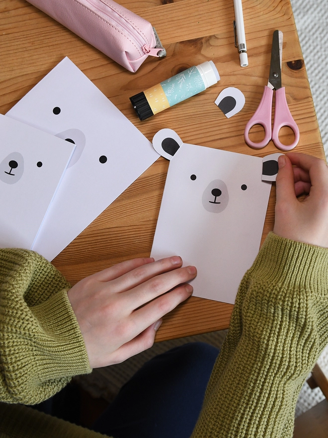 Several white polar bear greetings cards lay on a wooden table. A child is gluing the ears to the corners of one card.