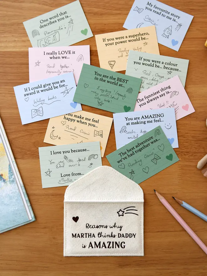 A flat lay of a handmade cream felt envelope with the words "Reasons why MARTHA thinks DADDY is AMAZING" on the front, surrounded by 12 colourful prompt cards filled in by a child with handwritten messages and drawings. The cards include prompts like "You are the BEST in the world at..." and "If I could give you an award...". Also visible are coloured pencils, a storybook, and a soft toy mouse in a tutu on a wooden desk.