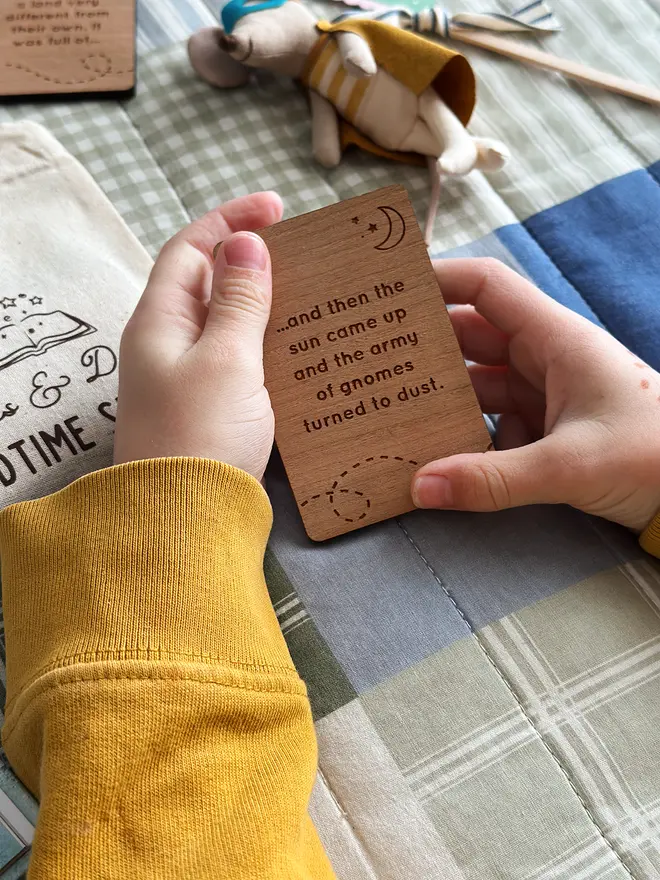 A child wearing a yellow jumper holds a wooden story card above a patchwork quilt.