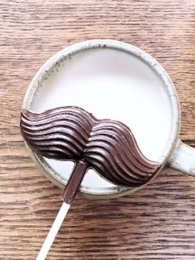 Moustache Chocolate Lolly 58%, a chocolate lolly sitting on a mug with a coffee in it. It sits on a wooden surface. 