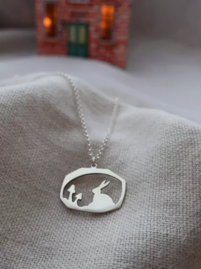 Silver Necklace rests on linen background, pendant shows sillouhette of rabbit and mushrooms, chain leads away into distance, background has dollhouse illumintated by candle, sterling silver, chain, childrens jewellery, heirloom gift
