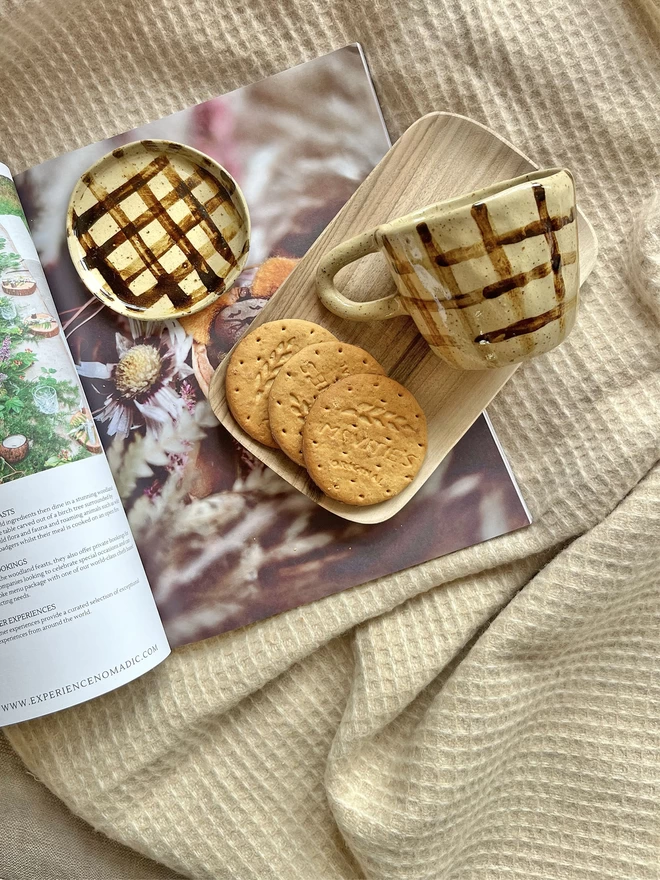 Beige stoneware mug and mini plate with a black chequered pattern laid on a wooden tray beside a pile of digestive biscuits