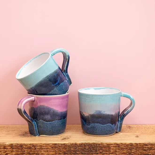 Landscape Standard Mug, a group of colourful mugs placed together on a wooden surface against a pink backdrop. 