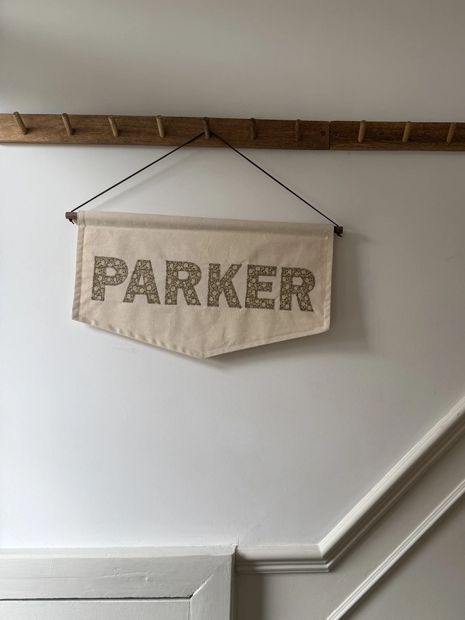 Personalised Fabric Name Banner. A large banner with PARKER in green floral fabric hanging from a peg rail. 