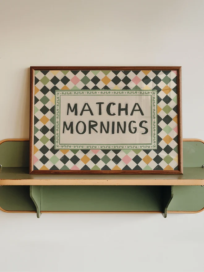 Matcha mornings checkerboard art prints on a wooden shelf on a white and green wall. 