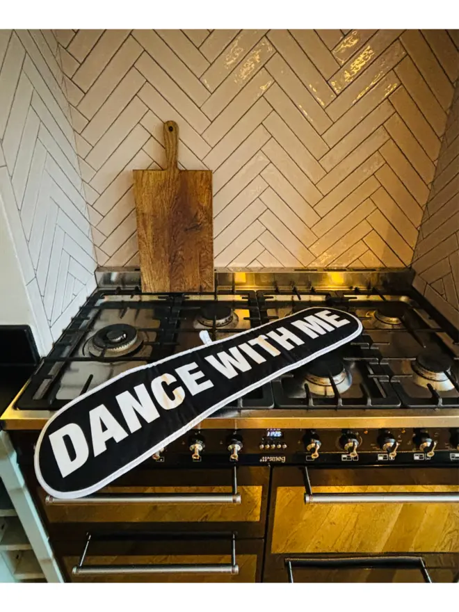 Black oven glove that says DANCE WITH ME on it laying on a hob with tiled wall and chopping board in background