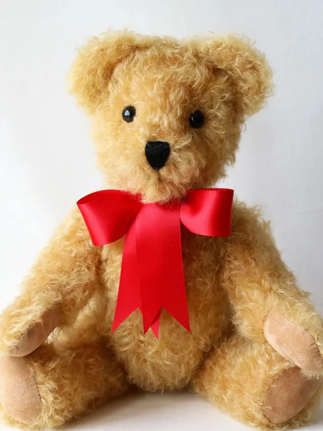 Handmade brown teddy bear sitting upright with a red ribbon bow against a plain background.