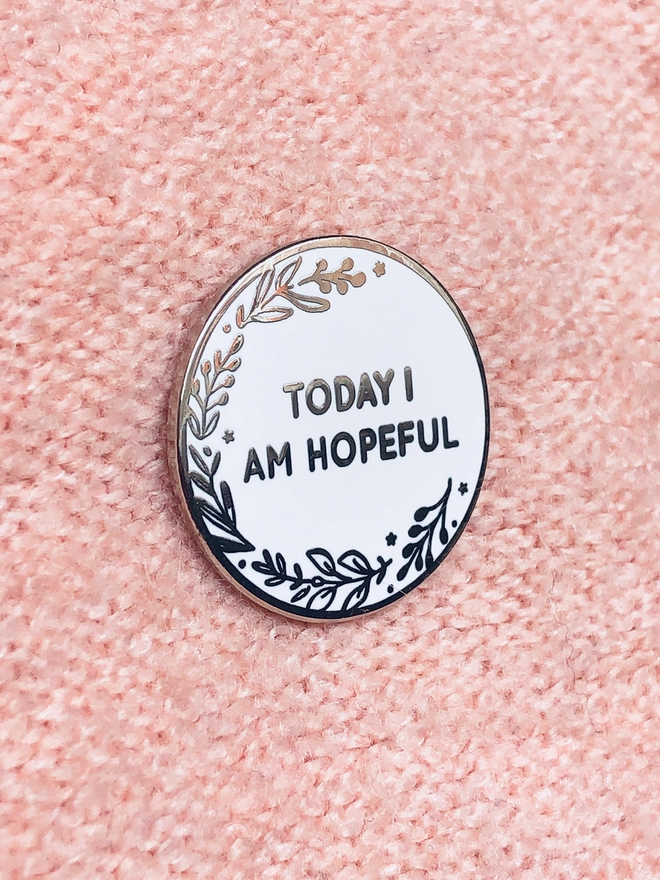 A round white enamel pin with a floral design and the words "Today I Am Hopeful" is pinned to pink fabric.