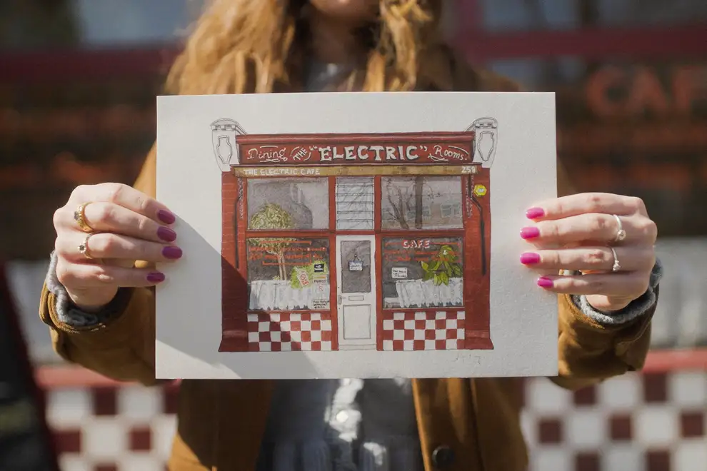 West Norwood The Electric Cafe Giclée Print Tulse Hill, a woman holding a print of a cafe in front of her. 