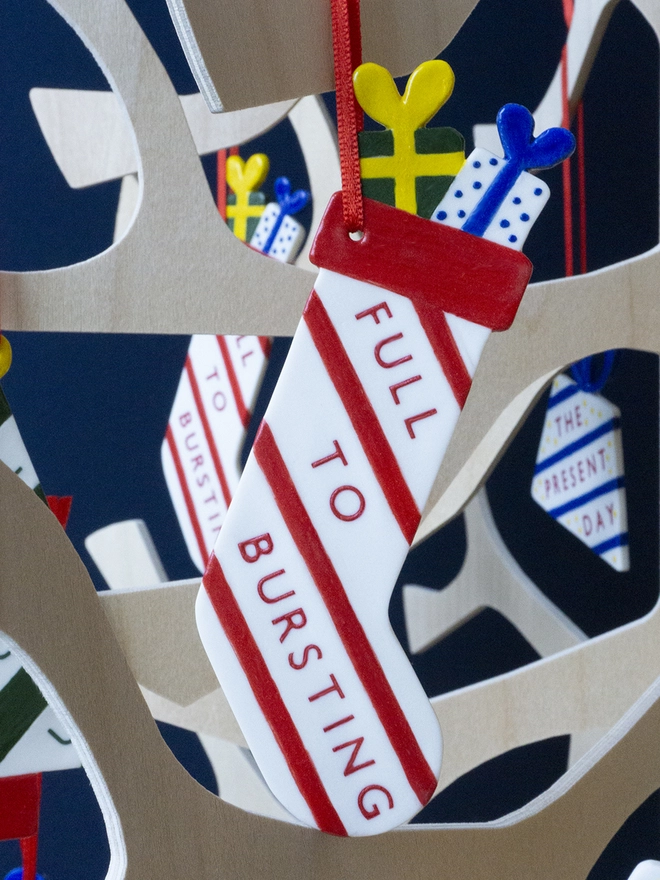 Ceramic porcelain Christmas decoration with Full to Bursting written on the side. A white stocking-shaped Christmas decoration with red lettering, decorated with red stripes and hanging from a plywood Christmas tree.