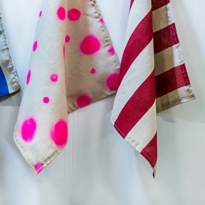 an offwhite cotton napkin, decorated with neon pink spots, hanging next to a cotton and red striped napkin, against a white backdrop