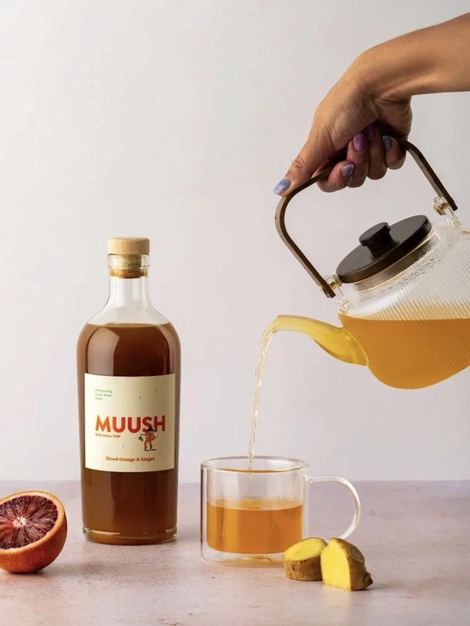 Blood Orange & Ginger Elixir, a drink being poured on a plain surface. It is accompanied by fruit. 