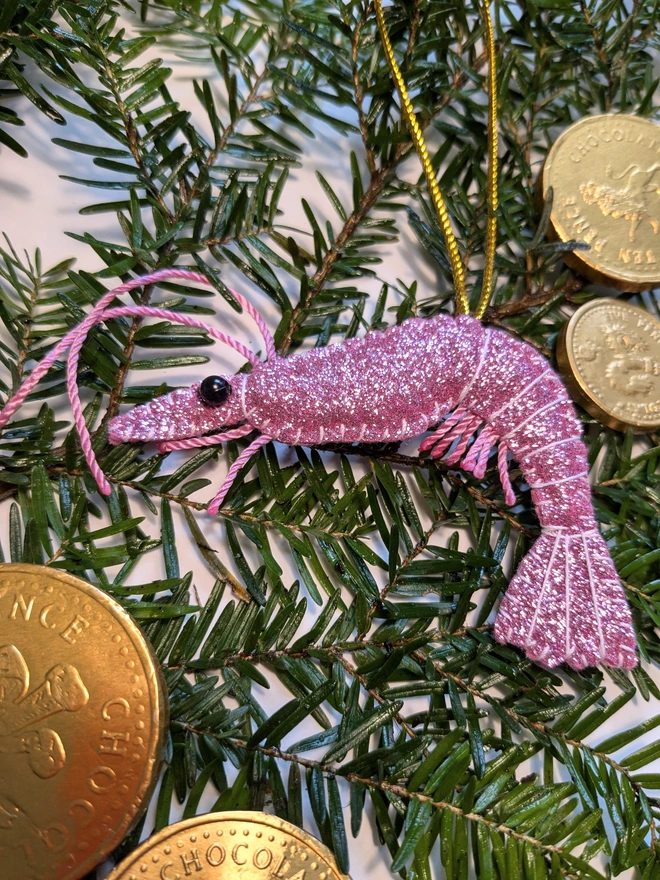 Hand-stitched pink glitter King Prawn Christmas decoration with pink twine antennae and legs, black beady eyes and gold hanging loop, nestled amongst festive greenery and gold chocolate coins.