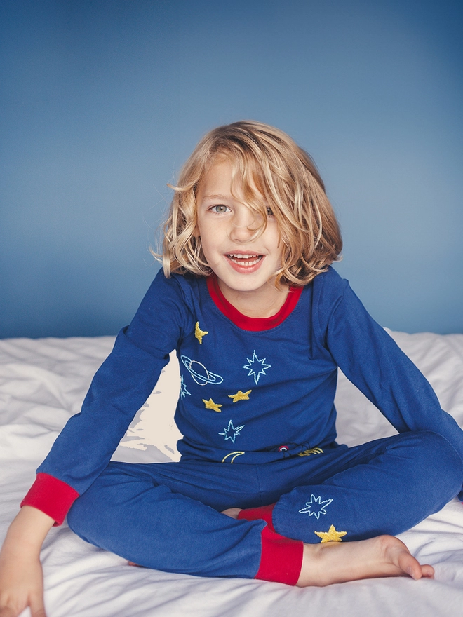 A boy in navy and red pjs with a space scene sits cross legged on a bed