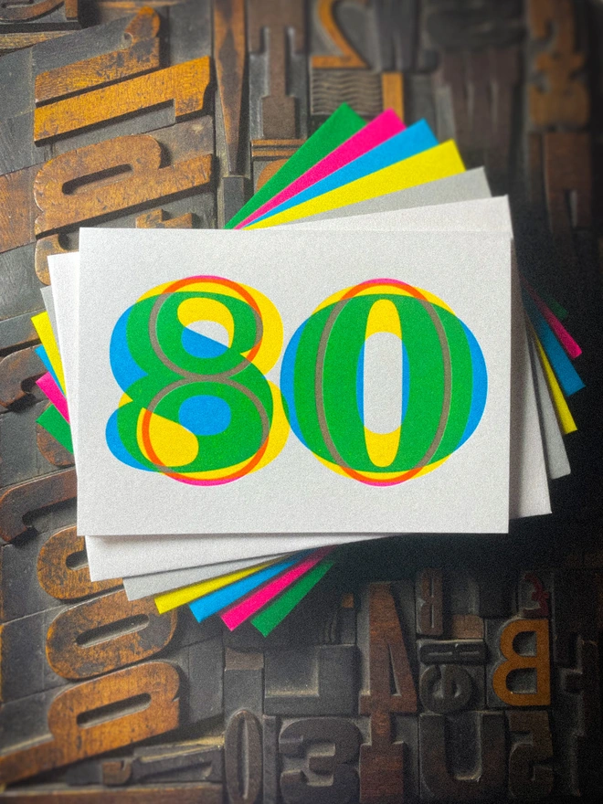 Handmade letterpress 80th birthday card featuring bold overlapping numerals printed in vibrant fluorescent cyan, magenta and yellow inks. Each card is individually hand printed using traditional letterpress techniques on luxury paper stock, creating a beautifully tactile finish with a subtle deboss and rich layered colours inspired by the CMYK printing process.