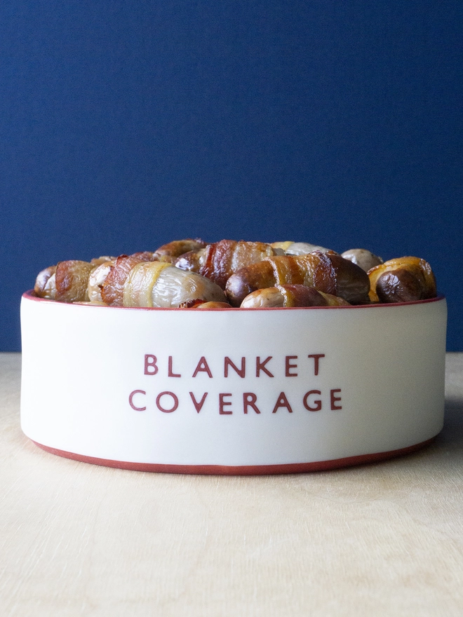 Ceramic porcelain dish with Blanket Covering written on the side. A white Christmas serving dish with red rim containing pigs in blankets.