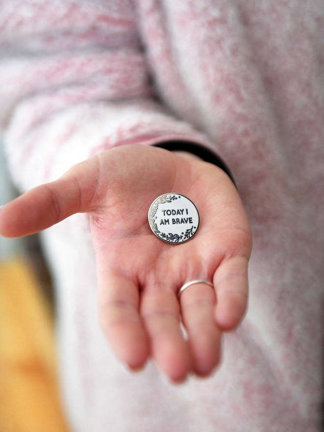 A round white enamel pin with a floral design and the words "Today I Am Brave" rests on the palm of a hand.
