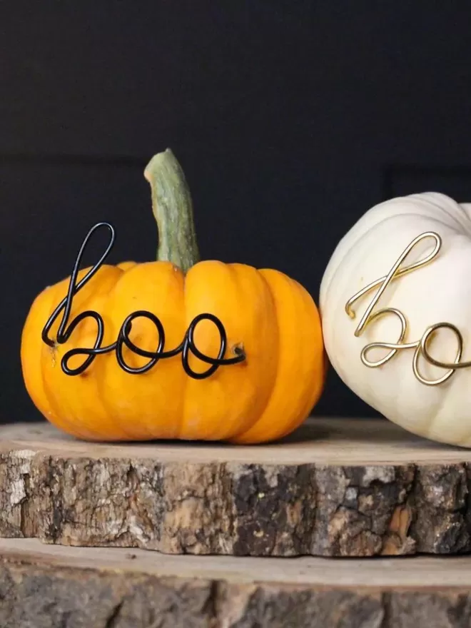 Mini boo in gold and black wire seen stuck into an orange and a white pumpkin for halloween.