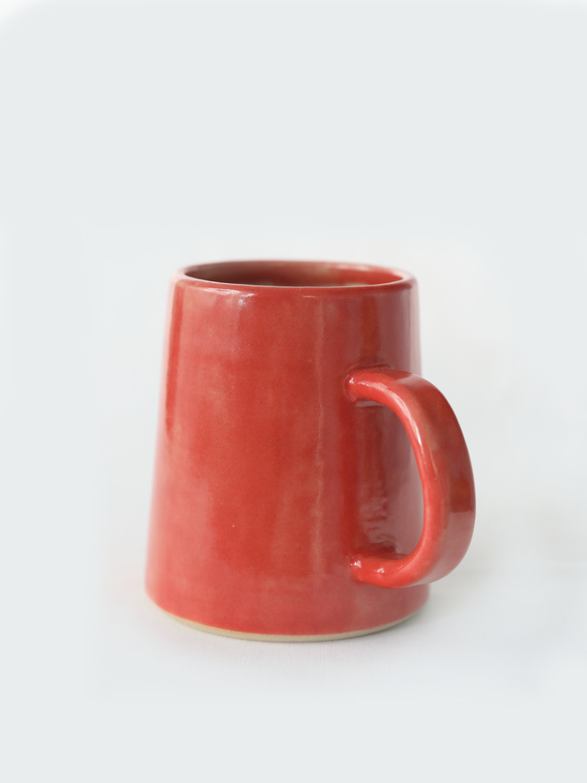 A handmade ceramic pint mug in cherry red glaze with a chunky loop handle, photographed against a plain light grey background