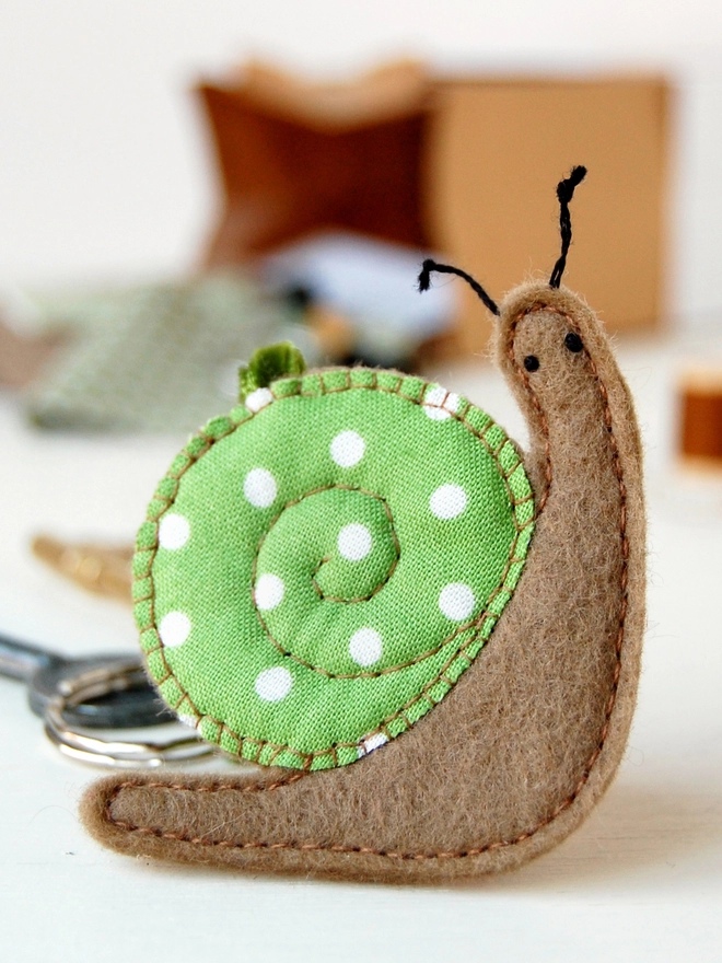 A homemade felt snail keyring with a green spotty cotton shell is on a white desk in front on the craft kit