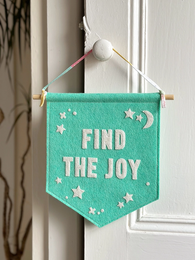 A turquoise felt banner with white words that read "Find The Joy" stitched on hangs from a ribbon hanger on a white wooden door.