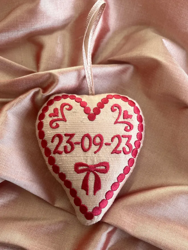 pink silk stuffed ornament with freehand embroidered design and date in red thread with pink metallic ribbon