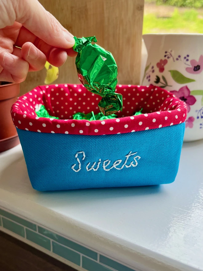 Blue fabric basket with red polka dot lining containing sweets with ‘Sweets’ embroidered on front.