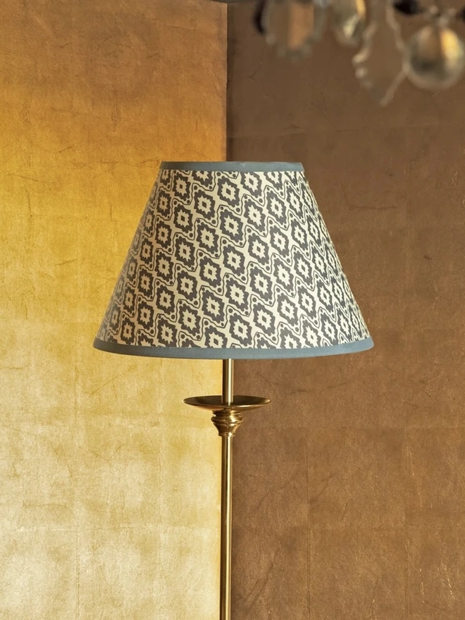 Paper Empire Lampshade. A tapered empire lampshade in grey and cream geometric ikat-style block print with a slate blue trim, fitted to a brass floor lamp. The patterned shade stands out against a warm gilded wall for an elegant living room lighting idea.