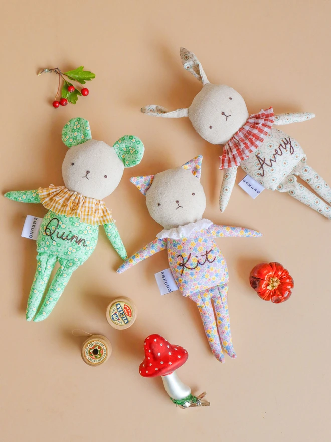 Three soft fabric dolls, one shaped like a mouse with round ears and a green floral outfit, one shaped like a cat with pointy ears and a pastel floral outfit and the other two shaped like a rabbit with long ears and a floral outfits, each with simple stitched facial features and a small “BOKUNO” tag on the side.