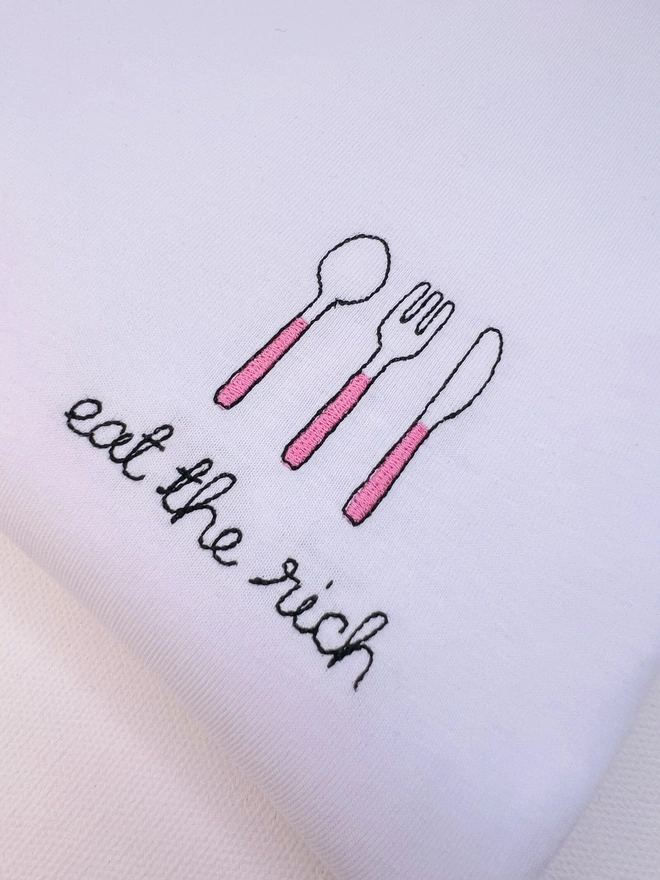     Close up of white T-shirt embroidered with pink cutlery motif and the words “Eat the Rich”
