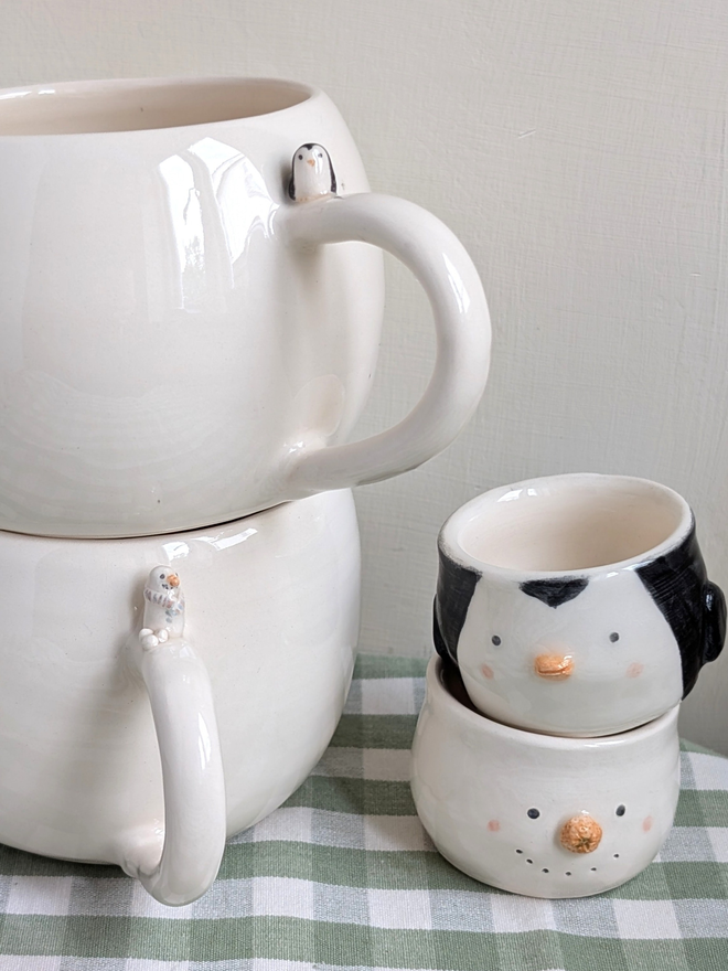 stack of 2 white cups with a tiny ceramic snowman and snowballs on the handle of one cup and a tiny penguin and bird prints on the other mug