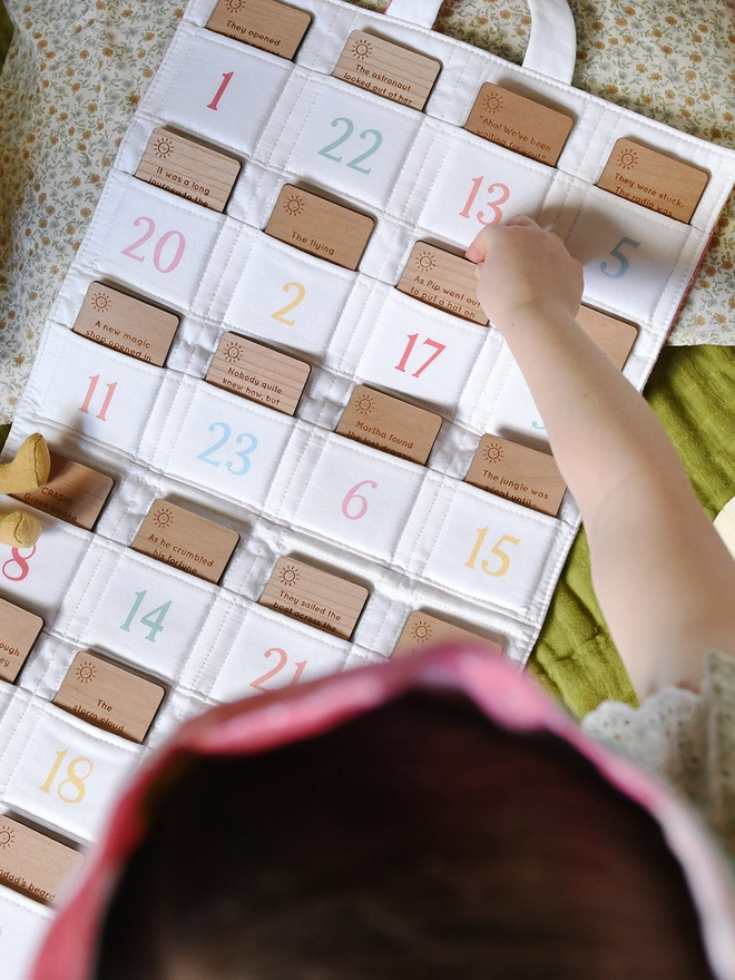 A fabric advent calendar with numbered pockets has a wooden story idea card tucked into each pocket. A child's hand is reaching for one of the cards.