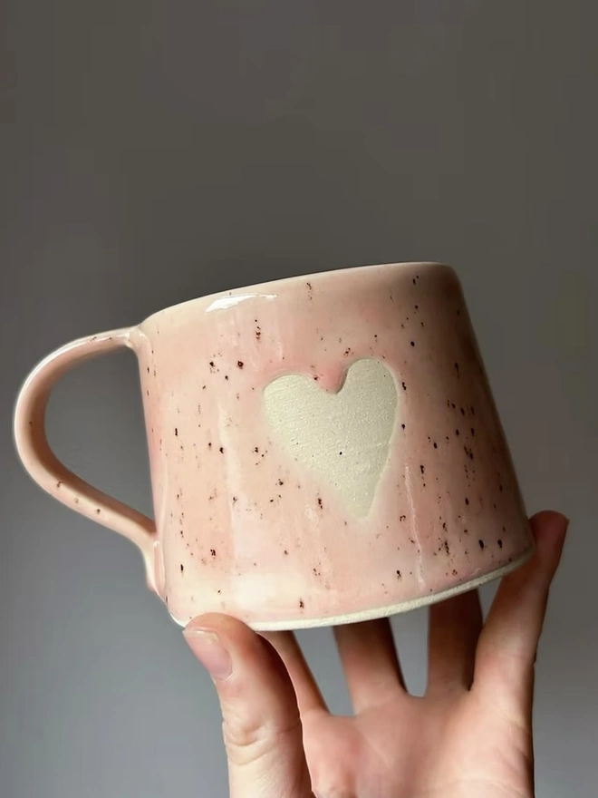 Heart Mug Strawberry Pink, person holding the base of the pink mug in front of a plain background. 