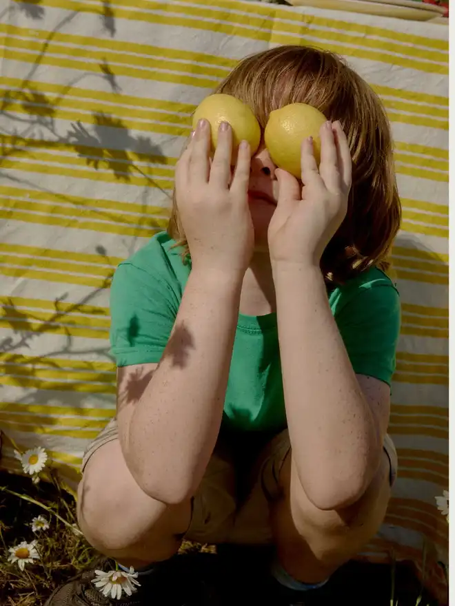little boy holding lemons infront of hi eyes sitting in front of yellow striped tablecloth