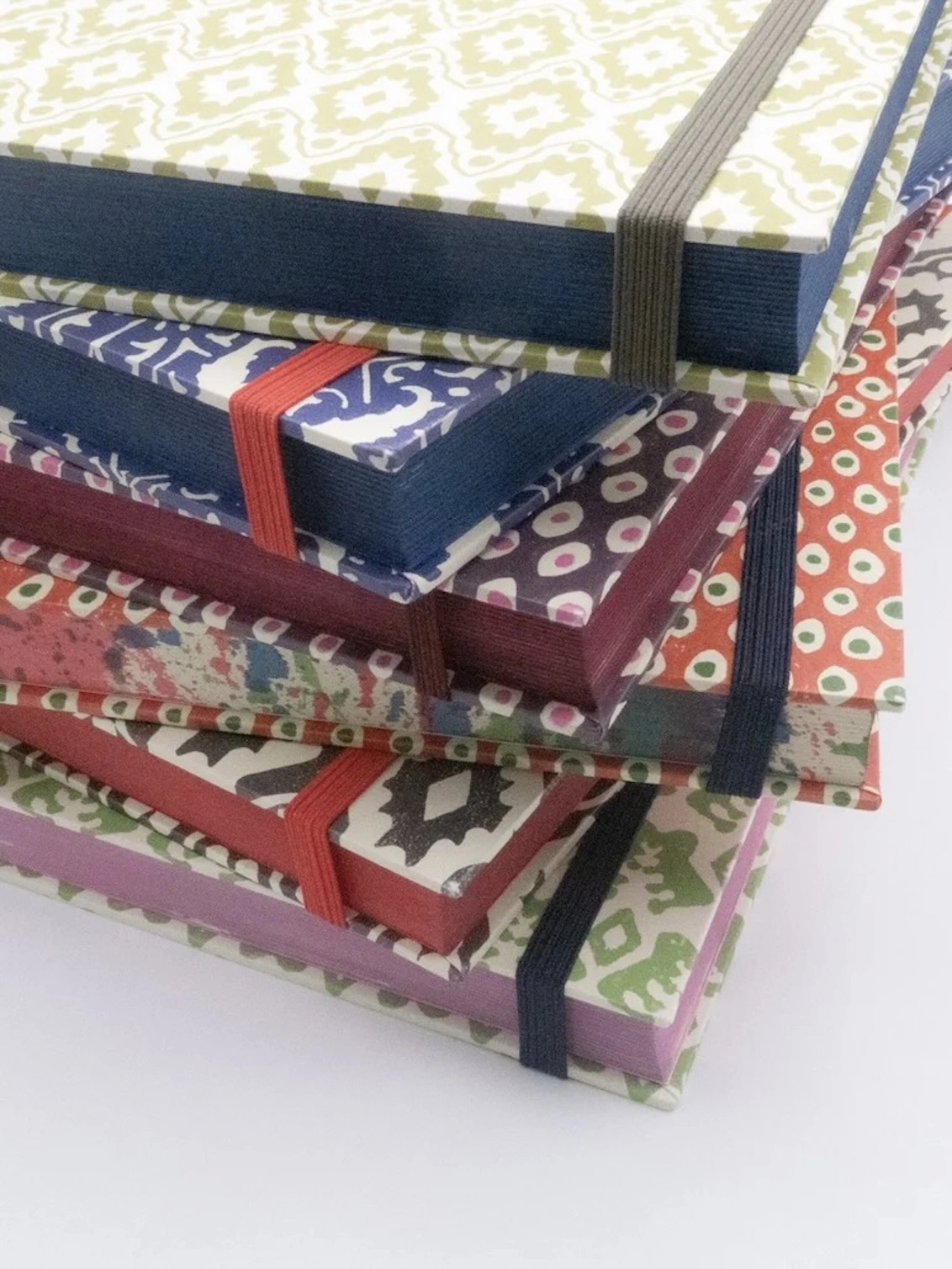 Large Sketch Book. A stack of hand block printed hardback notebooks in green, navy, plum, coral and charcoal patterns with coloured page edges. Each journal is fitted with a contrasting elastic closure for a vibrant, giftable stationery collection.
