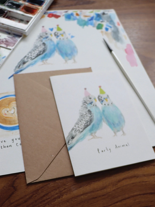 Close Up Slightly Out Of Focus Shot Of The Party Budgies Greeting Card Sitting Along Side The Original Hand Painted Watercolour Illustration, Paintbrush and Palette