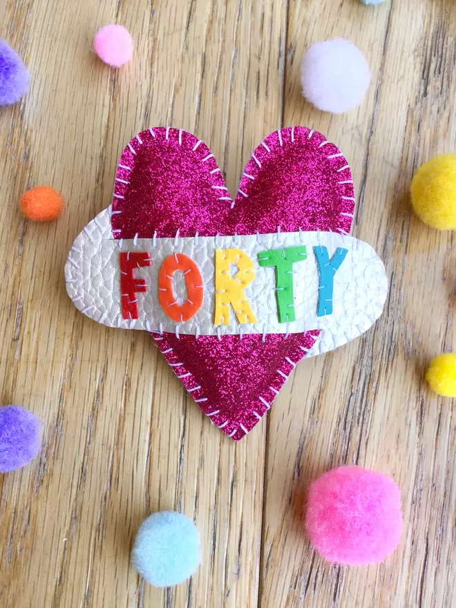 Pink glitter heart shaped brooch, with rainbow lettering spelling out FORTY on a white scroll, placed on a wooden surface, surrounded by pastel colour pom poms