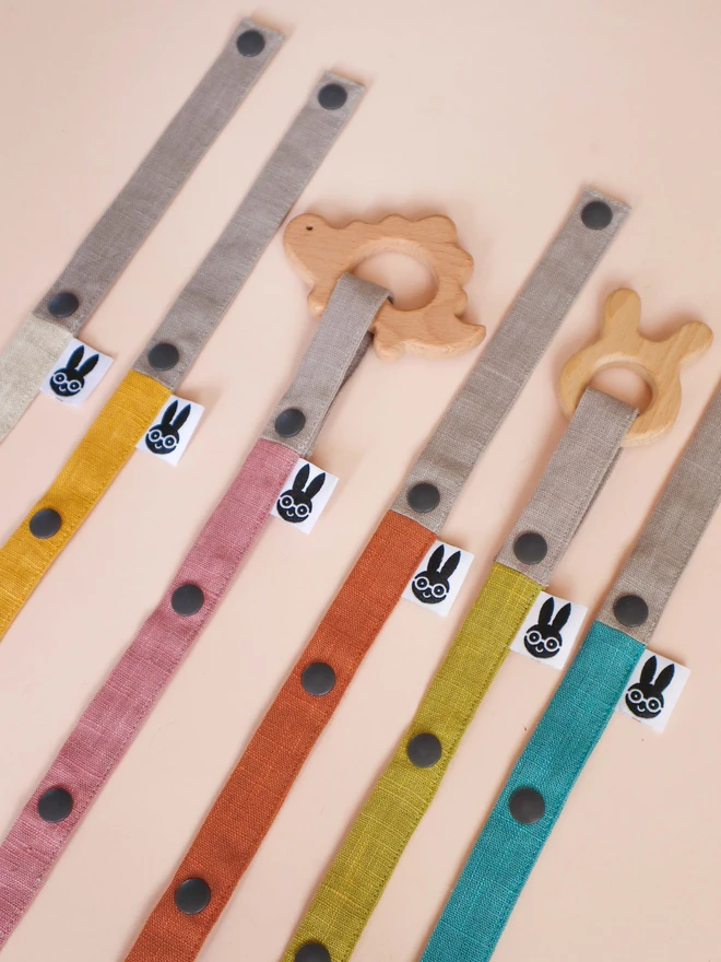 Handmade toy straps in soft neutral and pastel tones, featuring sturdy snap fastenings and small fabric tags with a bunny logo, neatly arranged in a row.