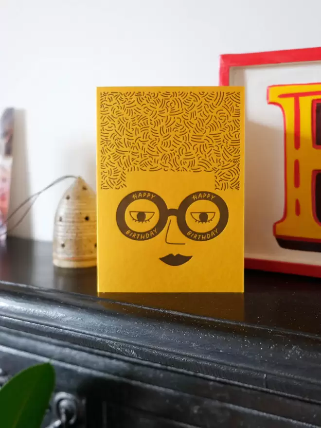 Yellow card with a ladies face wearing glasses saying ' Happy Birthday' and big square hair. 