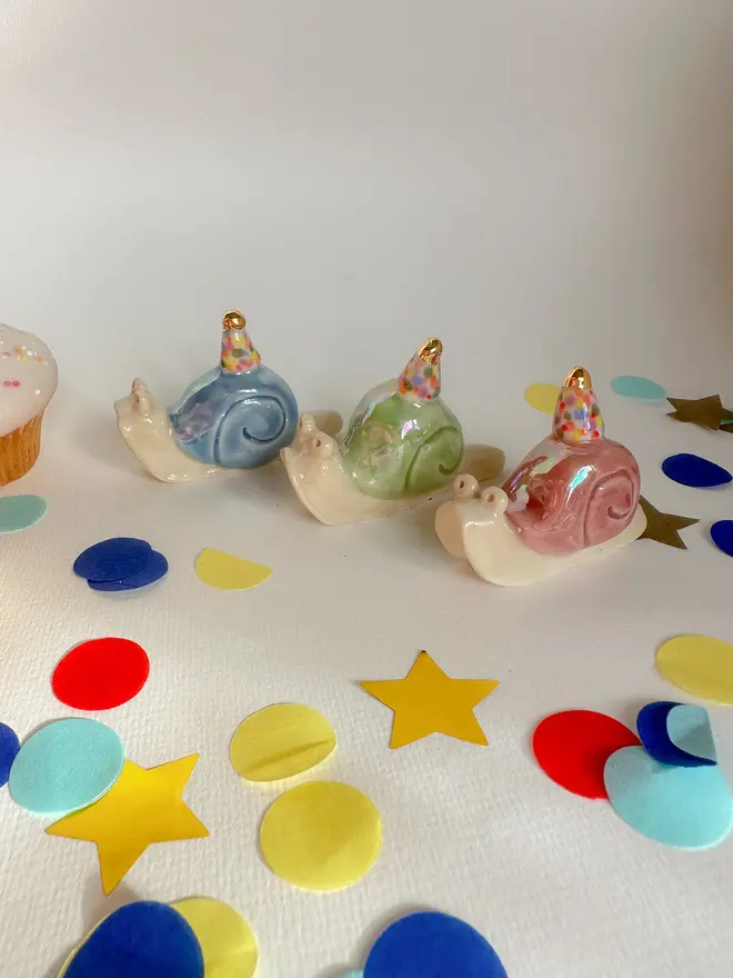 Handmade Porcelain Snail Cake Topper With Golden Hat, grouped together on a table with party decorations. 