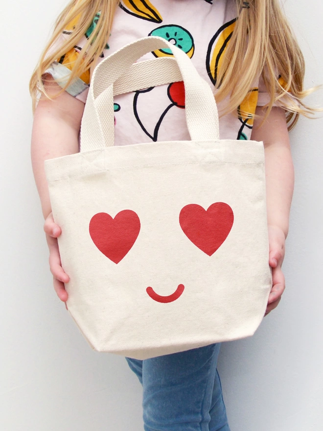 a child holding a mini kid's size tote bag with heart eyes and smiling mouth