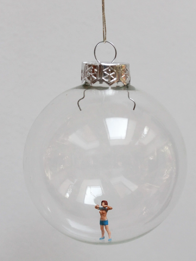 Miniature figure of a girl mischievously pulling up her top, in a glass hanging christmas bauble 