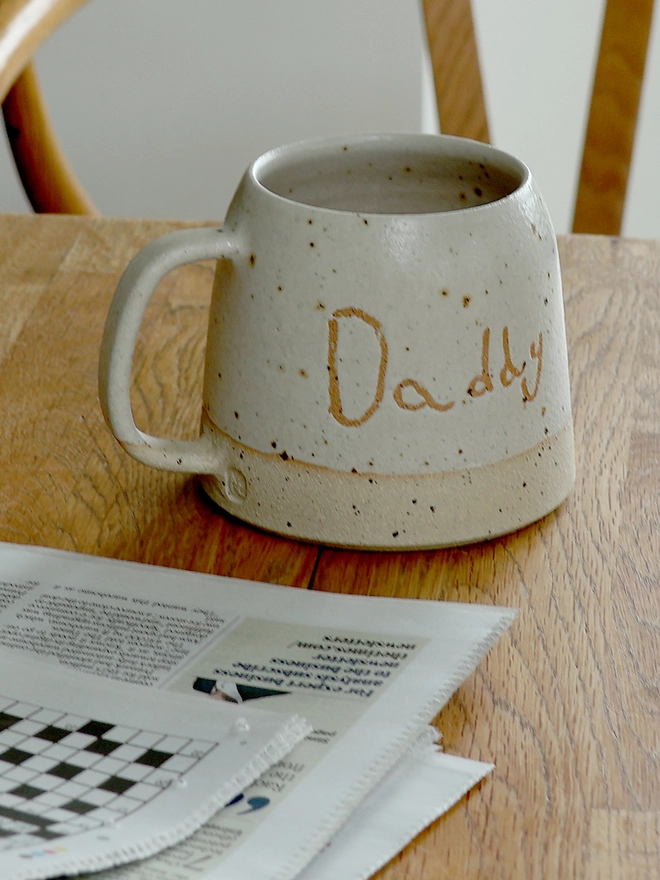 Personalised Kids' Handwriting Handmade Stoneware Mug with the name 'Daddy' beside a newspaper on a wooden table