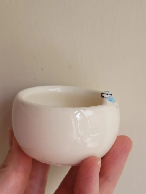 a small ceramic white pot is held in the finger tips of a hand the tealight holder has a tiny bluetit bird attached to the rim