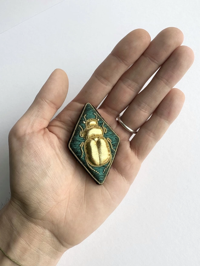Diamond shaped brooch with golden beetle on a green background sat in the palm of a hand 