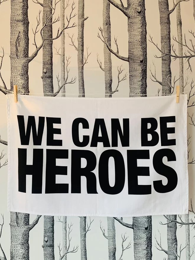 We Can Be Heroes printed on white tea towel hanging washing line style in front of wallpaper that is white with pattern of silver birch trees in black