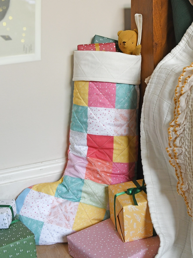 A quilted patchwork stocking in various pastel colours stands beside a wooden bed with ivory bedding. Small gifts and toys poke out of the top.