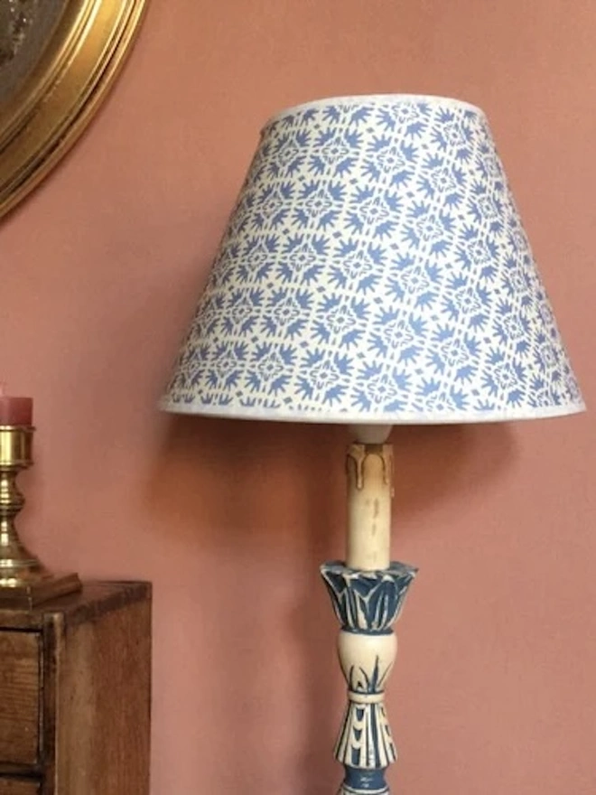 Paper Empire Lampshade. A tapered lampshade in a soft blue and cream floral block print fitted to a painted candlestick lamp against a dusky pink wall. The traditional patterned shade brings a fresh, country house feel to a bedroom or hallway.