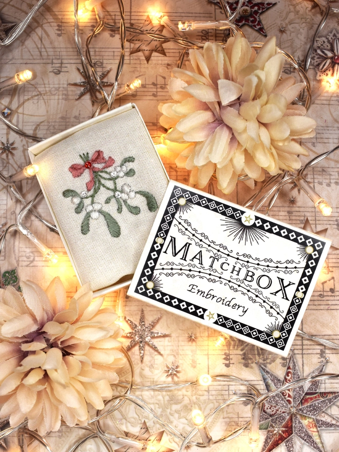 A sprig of mistletoe displayed in its matchbox.  surrounded by faux flowers and fairy lights.