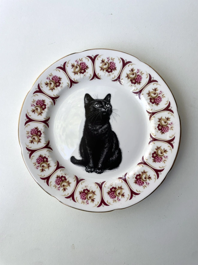 a vintage plate with an ornate border, in the centre of the plate is a vintage style black and white illustration of a black witch's cat